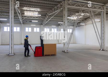 Jeune homme caucasien en casque, ouvrier dans l'entrepôt transportant la première palette à vide hall métallique. Stock de colis avec produits, conc. Transport Banque D'Images