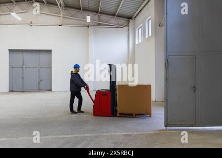 Jeune homme caucasien au casque, travailleur dans l'entrepôt transportant la première palette par la porte à la salle vide. Stock de colis avec des produits, transportati Banque D'Images