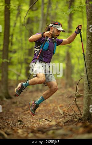 Un homme dans une chemise violette et un short gris saute par-dessus un arbre tout en tenant deux bâtons. Concept d'aventure et d'athlétisme Banque D'Images