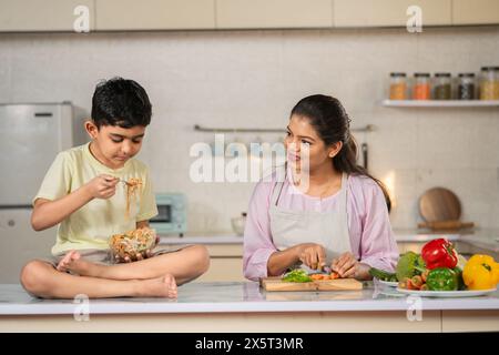 Enfant joyeux mangeant des nouilles à la cuisine tout en passant du temps avec la cuisine de la jeune mère de travail - concept de lien d'enfance, de parentalité et de santé Banque D'Images