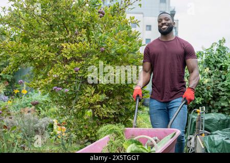 Portrait d'homme souriant avec des légumes dans la brouette dans le jardin urbain Banque D'Images