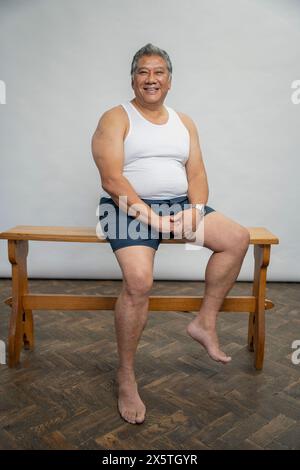 Portrait d'homme senior souriant assis sur un banc en bois sur fond gris Banque D'Images