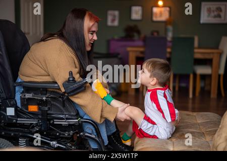 Femme en fauteuil roulant mettant sur la chaussure de football sur le pied fils dans le salon Banque D'Images