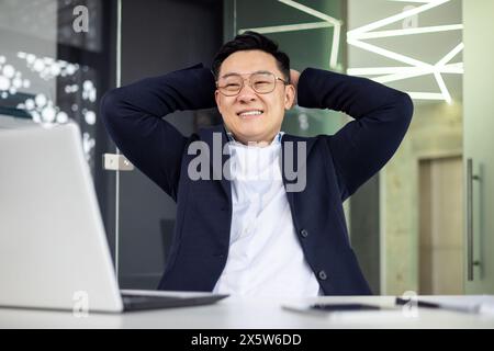 Joyeux homme d'affaires asiatique mature se penchant en arrière dans sa chaise de bureau, célébrant le succès avec un grand sourire dans un cadre de bureau moderne. Banque D'Images