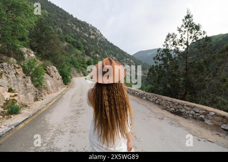 Une femme portant un chapeau marron marche sur une route. La route est vide et le ciel est nuageux. Banque D'Images