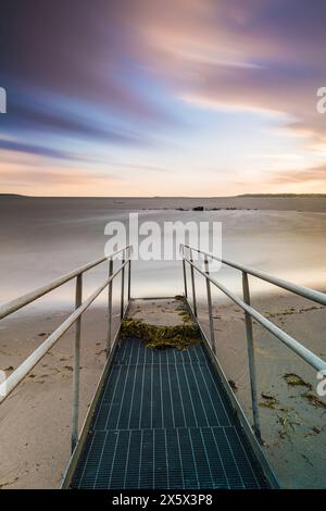 Une longue photo d'exposition d'une passerelle en béton s'étendant vers la vaste étendue de l'océan. La passerelle est éclairée par des lumières douces, créant un Banque D'Images