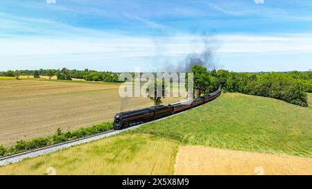 Vue aérienne captivante d'un train à vapeur vintage voyageant à travers un vaste paysage rural, avec des champs labourés et verts sous un ciel bleu. Banque D'Images