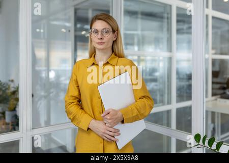 Entrepreneure confiante tenant un ordinateur portable, debout dans un bureau moderne lumineux avec une expression sereine, vêtue d'une chemise jaune élégante. Banque D'Images