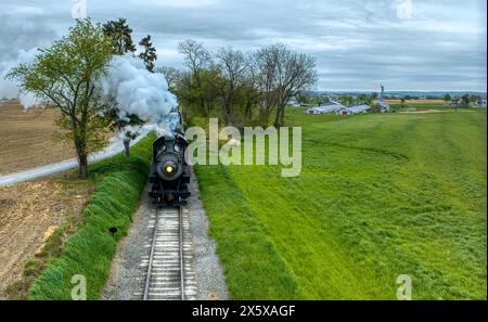 Plan atmosphérique d'un train à vapeur vintage traversant un paysage rural, émettant une épaisse fumée, avec en toile de fond des champs fraîchement labourés et une végétation luxuriante. Banque D'Images