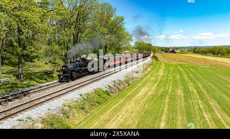 Une vue aérienne enchanteresse d'un train à vapeur historique émettant un nuage de fumée alors qu'il voyage à travers une campagne luxuriante, flanquée d'un champ vert vibrant Banque D'Images