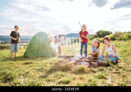 Trois sœurs rient et rôtisent gaiement des guimauves et des bonbons sur des bâtons au-dessus de la flamme de feu de camp tandis que deux frères dressent la tente verte. Heureux Banque D'Images