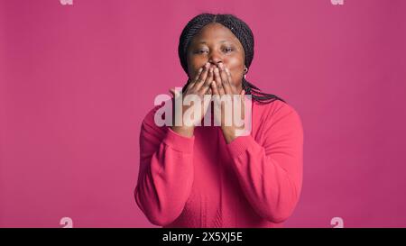 Dame à l'origine ethnique afro-américaine soufflant un baiser vers la caméra étant belle isolée sur fond rose. Superbe femme noire joyeuse exprimant l'amour avec de mignons gestes de la main embrassant. Banque D'Images