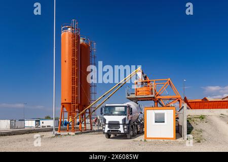 Cimenterie industrielle orange vif avec silos de stockage, entourée d'un sol en béton contre un ciel bleu vif Banque D'Images