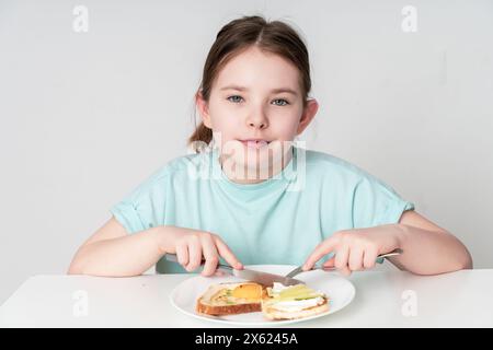 Une petite fille mignonne prend le petit déjeuner avec bruschetta avec oeuf, concombre et fromage cottage. Adolescente mangeant un petit déjeuner sain sur fond blanc. Le Banque D'Images