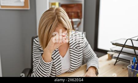 Femme blonde mature dans l'intérieur du bureau regardant stressé et fatigué tout en tenant ses lunettes. Banque D'Images