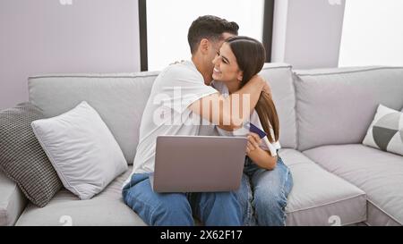 Un couple aimant câlin sur un canapé dans un salon moderne, avec la femme tenant une carte de crédit et un ordinateur portable en arrière-plan. Banque D'Images