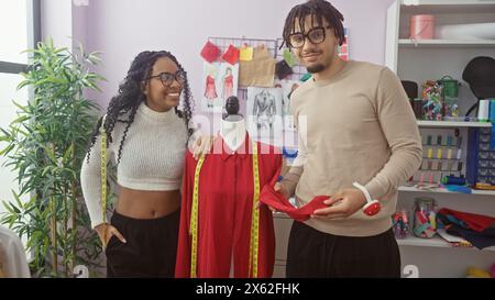 Une femme et un homme créateurs de mode sourient à côté d'un mannequin dans un magasin de tailleur coloré avec des vêtements, du fil et des croquis. Banque D'Images