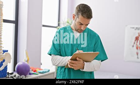 Bel homme hispanique en gommage écrivant des notes dans une salle de clinique de rééducation lumineuse. Banque D'Images