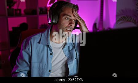 Homme hispanique avec des écouteurs dans une pièce sombre semble stressé tout en utilisant un ordinateur la nuit. Banque D'Images