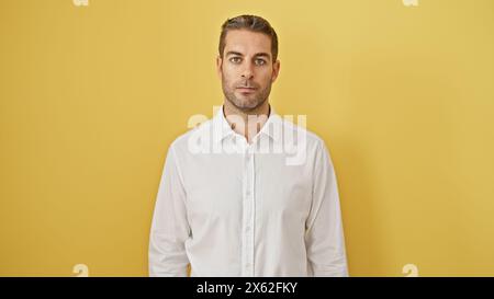 Bel homme hispanique barbu dans une chemise blanche posant avec confiance contre un mur jaune isolé Banque D'Images