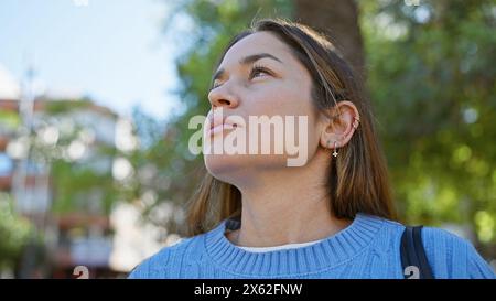 Une jeune femme contemplative aux cheveux longs bruns regarde vers le ciel à l'extérieur dans un parc ensoleillé, exsudant la paix. Banque D'Images
