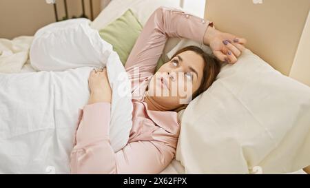 Une femme pensive se trouve dans son lit regardant loin, avec son pyjama élégant, son cadre de chambre confortable et son expression réfléchie. Banque D'Images