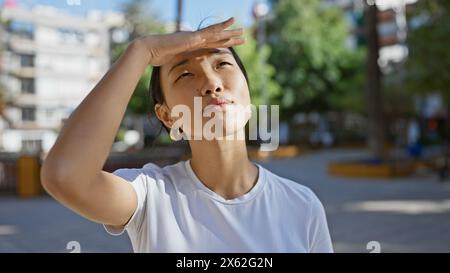 Une jeune femme asiatique en chemise blanche protège ses yeux du soleil dans un parc urbain verdoyant. Banque D'Images