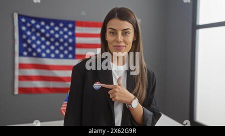 Une jeune femme hispanique confiante montre son autocollant « j'ai voté », mettant en valeur l'engagement civique sur fond de drapeau américain dans un intérieur Banque D'Images