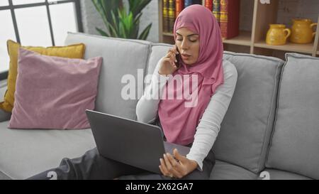 Une jeune femme dans un hijab engage une conversation au téléphone tout en travaillant sur son ordinateur portable dans un salon moderne. Banque D'Images