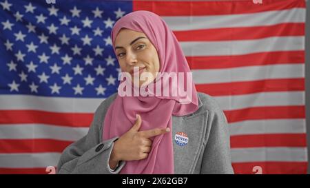 Une jeune femme confiante avec un hijab pointe vers son autocollant «j'ai voté» contre un drapeau américain en toile de fond. Banque D'Images