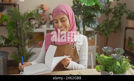 Une femme souriante dans un hijab écrivant à l'intérieur entouré de plantes d'intérieur, illustrant un environnement de maison confortable. Banque D'Images