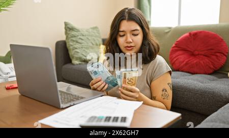 La jeune femme hispanique compte des reais brésiliens dans le salon d'un appartement confortable avec un ordinateur portable sur la table. Banque D'Images