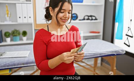 Une jeune femme hispanique souriante compte des reais brésiliens dans une salle de clinique thérapeutique, reflétant le bien-être et la finance de la santé. Banque D'Images