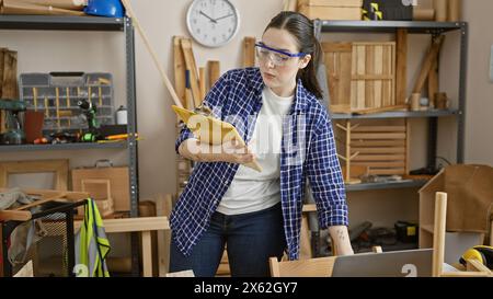 Femme caucasienne avec des lunettes de protection examine les plans dans un studio de menuiserie entouré d'outils Banque D'Images