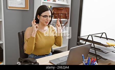 Une jeune femme hispanique pleine d'espoir croise les doigts pour porter chance tout en travaillant à son bureau, entourée de technologie et de fournitures de bureau. Banque D'Images