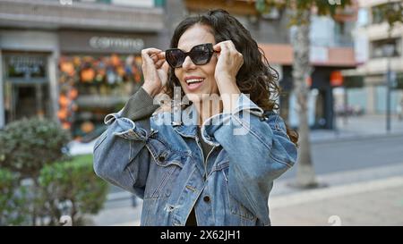 Femme hispanique d'âge moyen souriante avec les cheveux bouclés ajustant les lunettes de soleil sur une rue urbaine ensoleillée. Banque D'Images