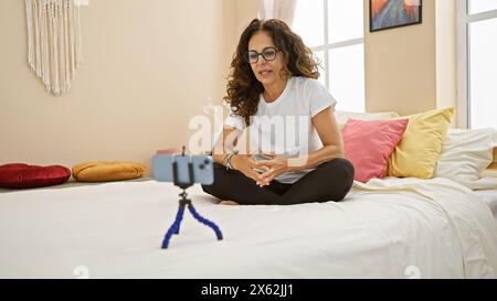 Femme hispanique mature vlogging dans la chambre avec des oreillers colorés, t-shirt blanc et lunettes. Banque D'Images