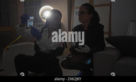 Deux enquêteurs femmes analysent les preuves à l'intérieur sur une scène de crime faiblement éclairée. Banque D'Images