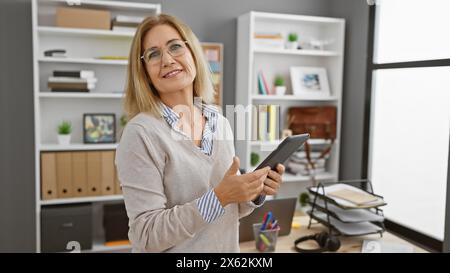 Une femme blonde, mature et souriante tenant une tablette dans un bureau moderne, représentant un adulte professionnel caucasien sur son lieu de travail. Banque D'Images