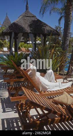 Une femme détendue se prélassant sur un transat en bois dans un luxueux complexe balinais, entouré de végétation tropicale. Banque D'Images