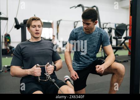 Portrait de la formation de sportif débutant dans le gymnase avec entraîneur personnel, expliquant comment travailler sur la machine à ramer. Homme faisant des exercices pour les muscles sous Banque D'Images