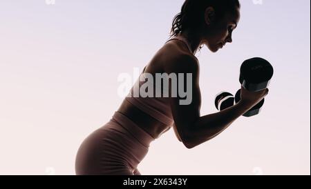 Fit femme dans les vêtements de sport soulève des haltères dans un studio de fitness. Sa routine de musculation se concentre sur le cardio et le renforcement musculaire. Silhouette d'un woma Banque D'Images