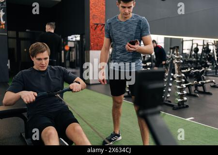 Jeune sportif débutant fatigué s'entrainant dans la salle de gym avec entraîneur personnel, expliquant comment travailler sur la machine à ramer. Homme faisant des exercices pour les muscles sous Banque D'Images