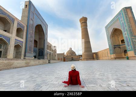 Femme touristique avec chapeau et robe rouge assis sur la place POI Kalyon une ancienne place publique au cœur de la ville antique de Boukhara, Ouzbékistan. Banque D'Images