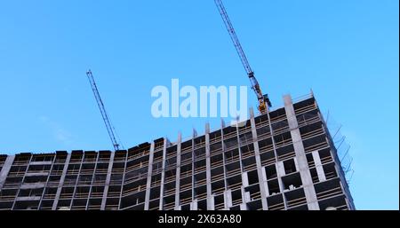 Deux grues de construction s'élèvent au-dessus d'un bâtiment de plusieurs étages sous construction en béton, les fenêtres n'ayant pas encore été installées, sur fond de ciel bleu. Idéal Banque D'Images