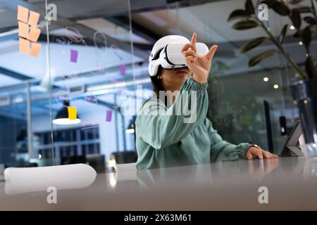 Travaillant tard au bureau, femme d'affaires biraciale portant un casque VR, pointant vers le haut Banque D'Images