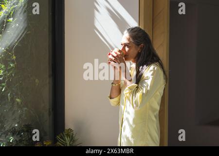 À la maison, femme caucasienne mature avec de longs cheveux gris, tenant la tasse par la fenêtre Banque D'Images