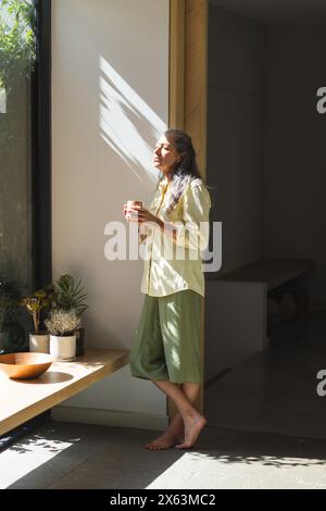 À la maison, femme caucasienne mature tenant une tasse par fenêtre Banque D'Images