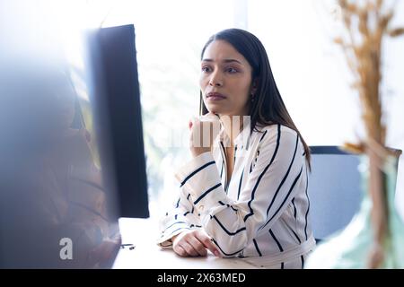 Femme biraciale d'âge moyen se concentre sur l'écran d'ordinateur, chemise rayée, au bureau Banque D'Images