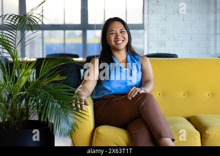 Au bureau, femme d'affaires biraciale assise sur un canapé jaune, souriante Banque D'Images
